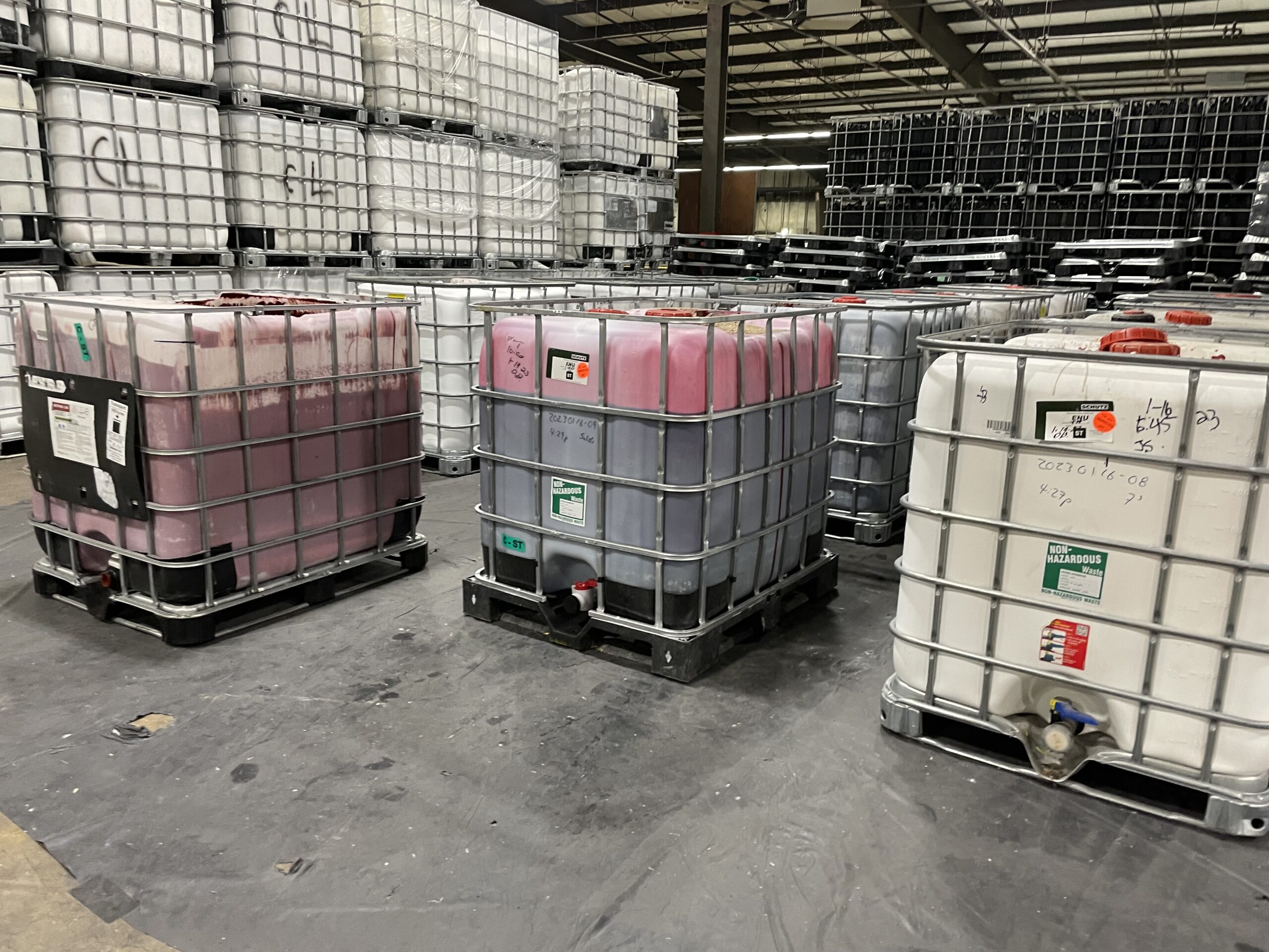 Photo of chemical totes encased in steel cages lined up in a large warehouse where facility auditing is conducted.