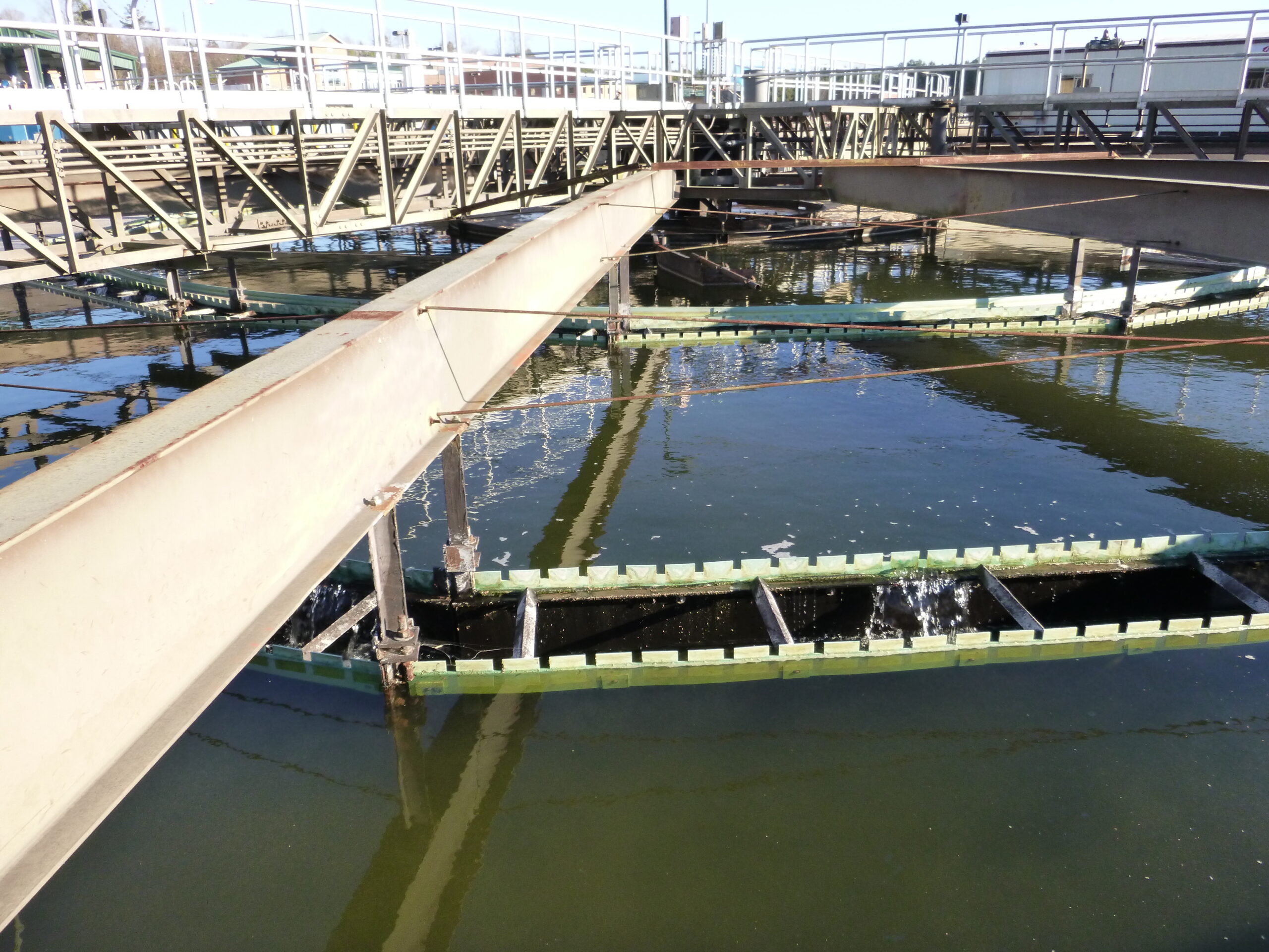 Photo of steel beams and walkways positioned over a dark body of water with a metallic green wastewater treatment unit. This unit is called a secondary clarifier and it separates biological solids, microbes and germs from treated liquid waste.