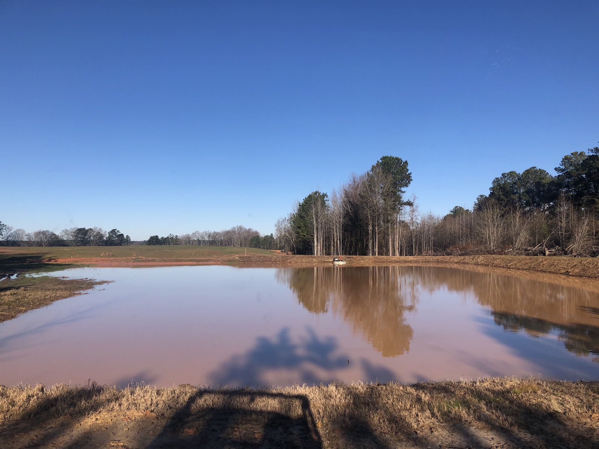 Photo of a wide, brown pond surrounded by brown grass in the foreground, trees in the background and a blue sky.