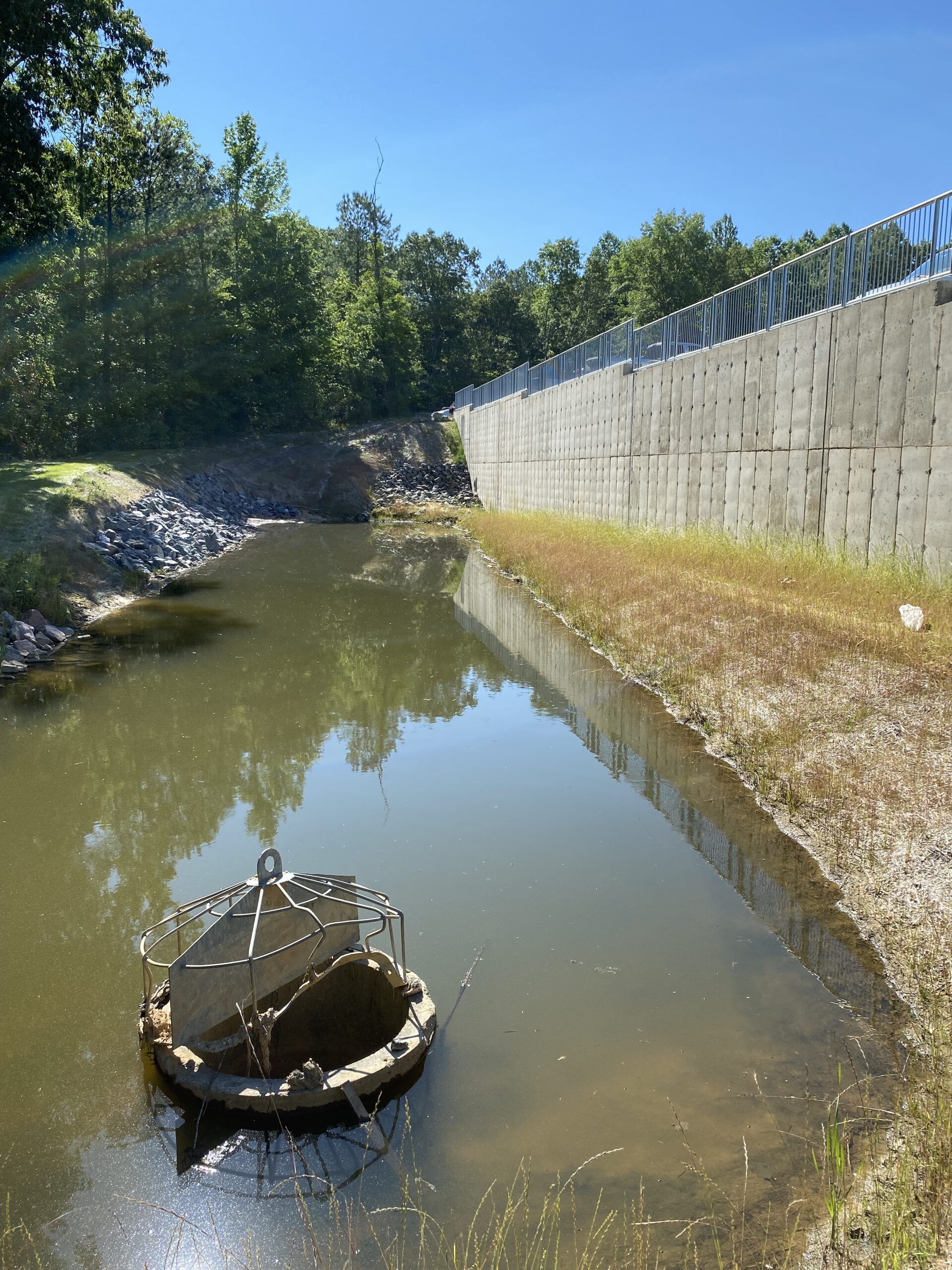 Photo of a retention pond with a metal drainage structure in the foreground, surrounded by sand, grass and rocks, and bordered by a concrete retention wall with metal fencing on top.