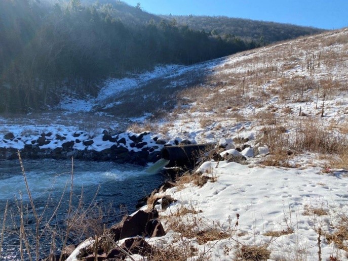 Snow-covered landscape with a flowing stream and hills in the background.