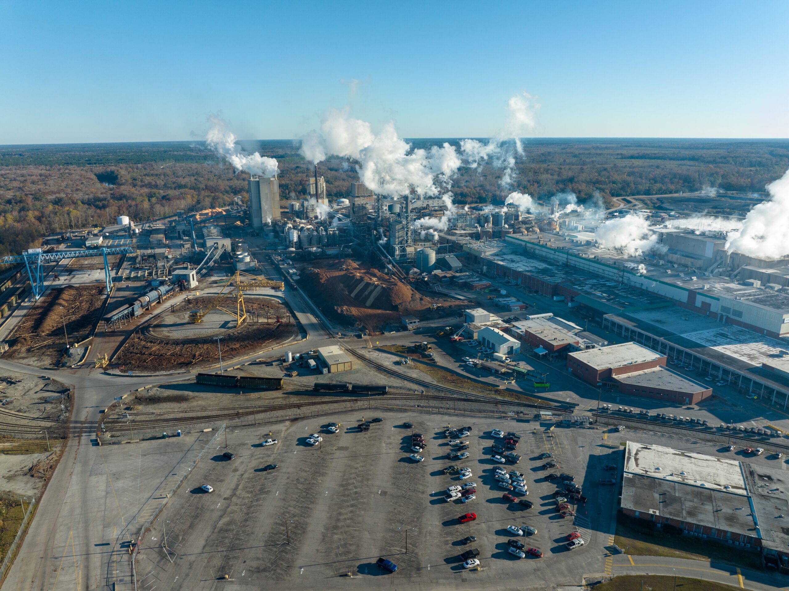 Aerial view of a large industrial complex with steam coming from pipes and a parking area below.