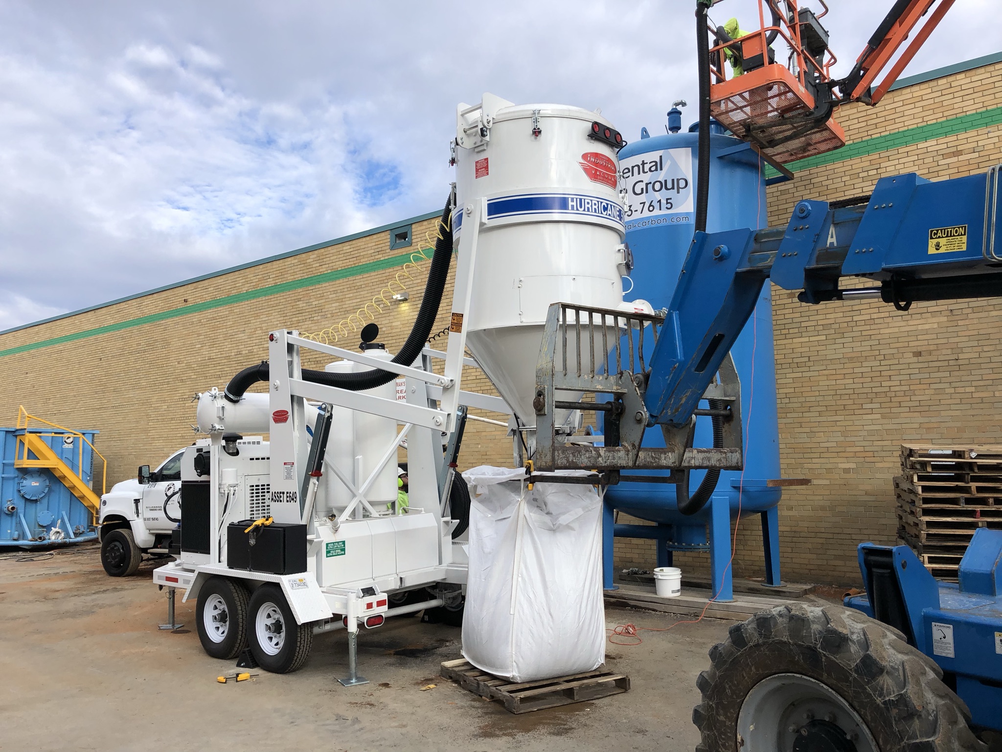 Photo of a large brick building with a blue tank and a white tank, orange lift, and other equipment working to funnel into a large, white bag.