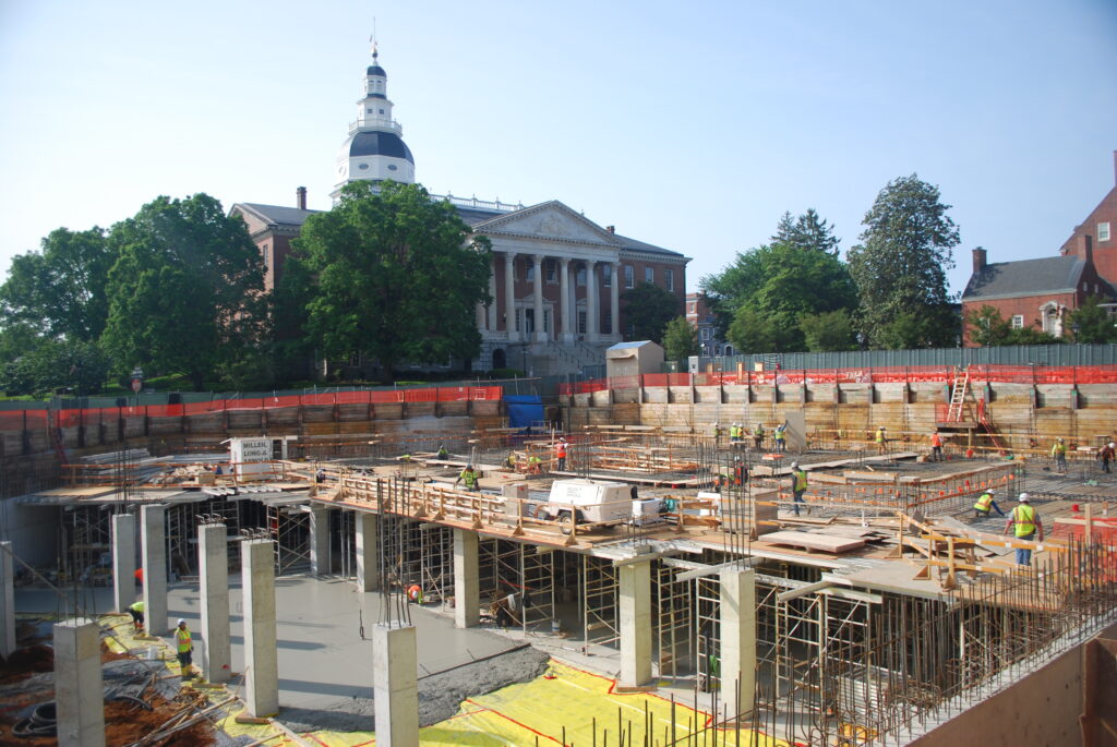 Photo of the Maryland State House surrounded by trees in the background. In the foreground is the foundation for a new building surrounded by orange safety fencing. People are working on the foundation.